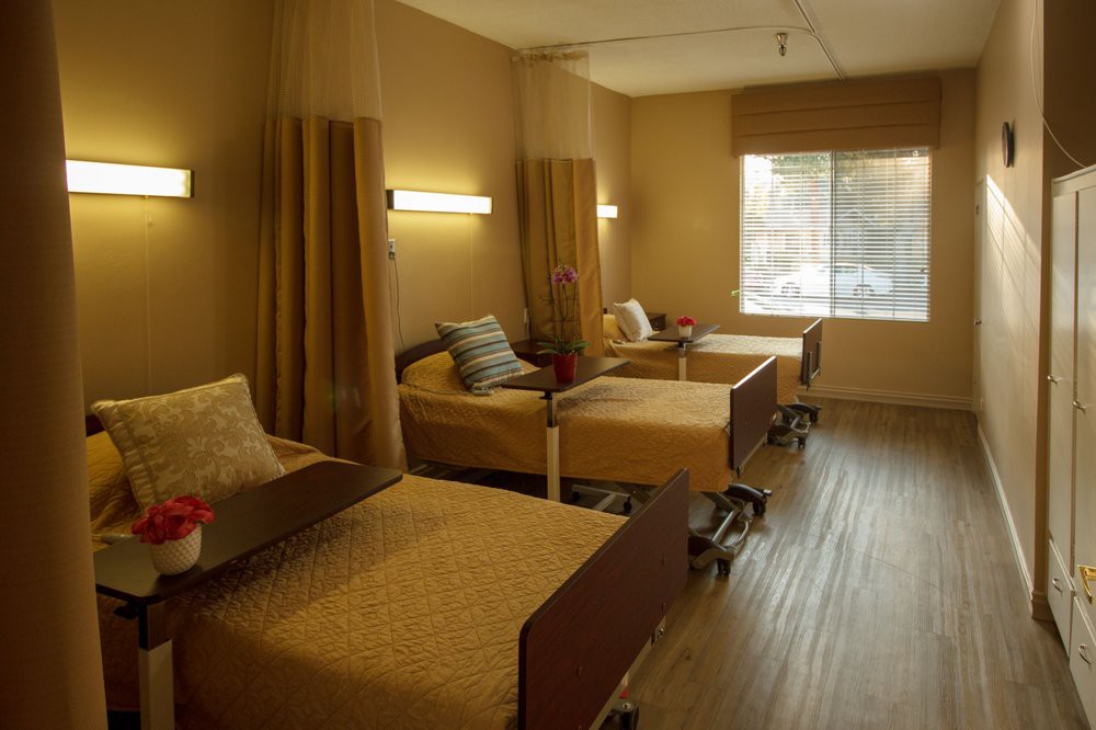 A shared healthcare room with two beds, separated by curtains and lit by wall-mounted lights.