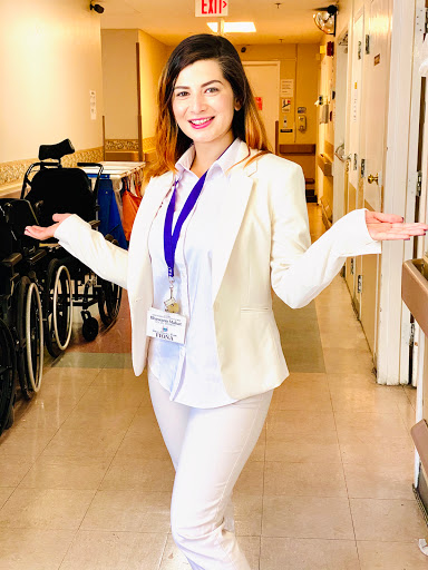A smiling woman in professional attire gestures welcomingly in a hospital setting.