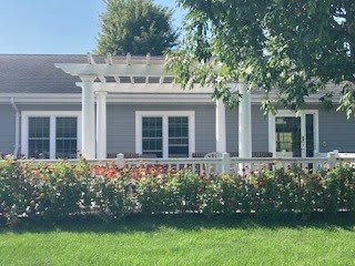A cozy house with a white pergola over the porch, surrounded by green hedges and a neatly maintained lawn.