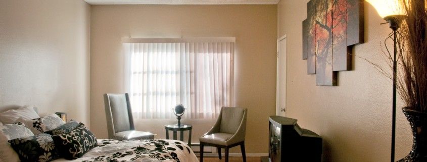 A cozy bedroom with two chairs, a small table, and a window with sheer curtains.