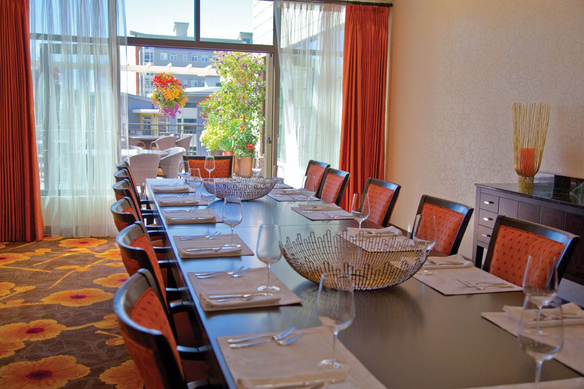 A well-set dining table in a bright room with large windows and orange curtains.