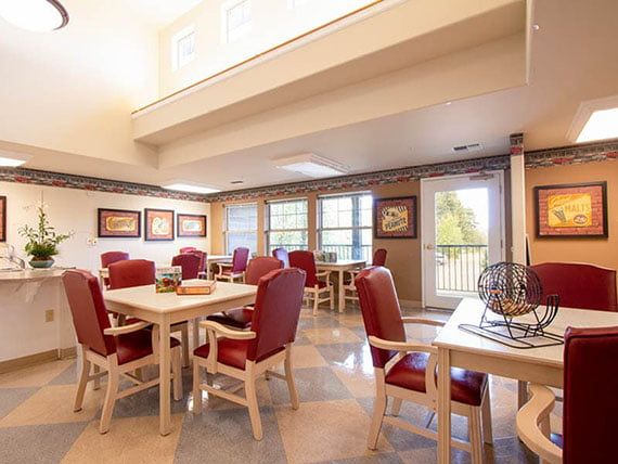 A spacious dining room with red upholstered chairs and wooden tables, well-lit by natural light streaming through large windows.