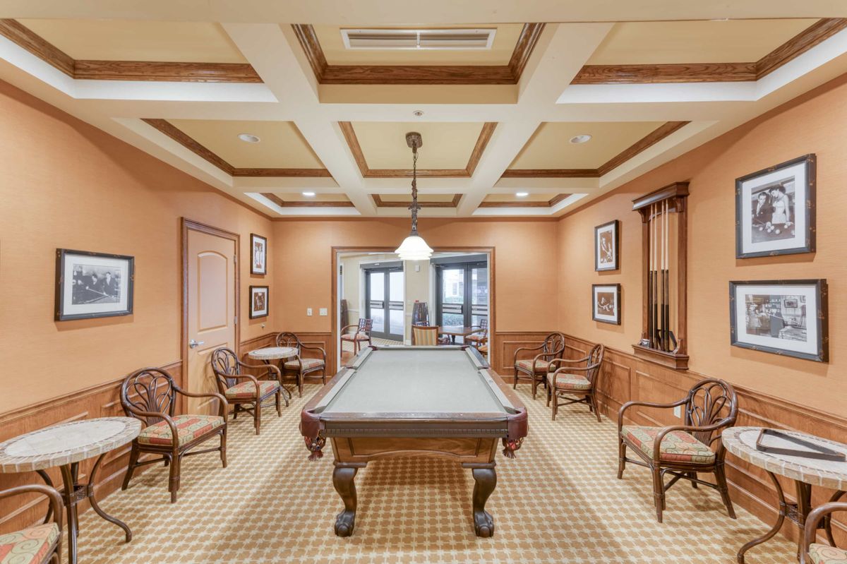 An elegantly designed billiards room with a central pool table, surrounded by classic wooden chairs and framed wall art.