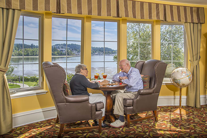 Two seniors enjoying a meal with a scenic lake view through large windows.