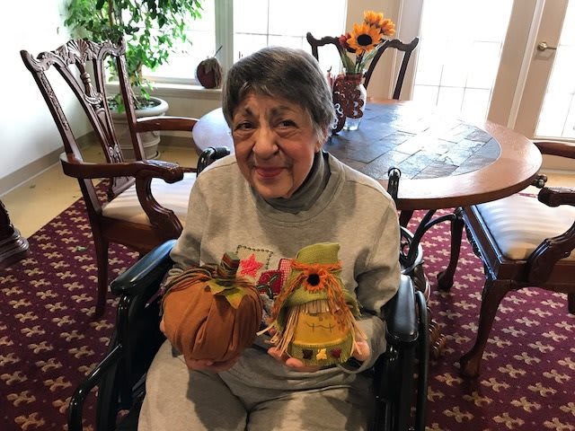 An elderly woman in a wheelchair smiles while holding crafts and a small pumpkin indoors.