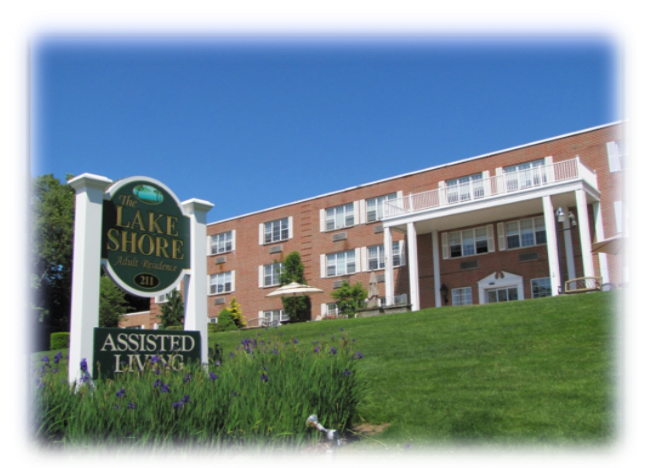 A sunny day view of an assisted living facility with a sign and landscaped front yard.