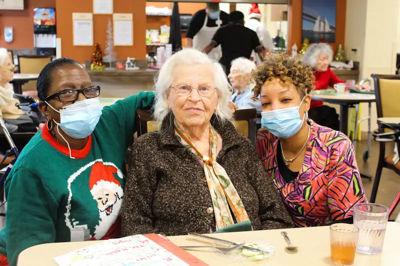 Three women, two wearing masks, gather together at a community event.
