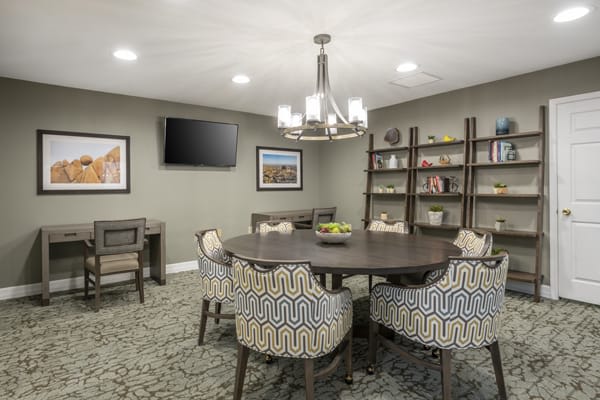 A modern conference room with a round table, patterned chairs, a chandelier, bookshelves, and wall-mounted television.