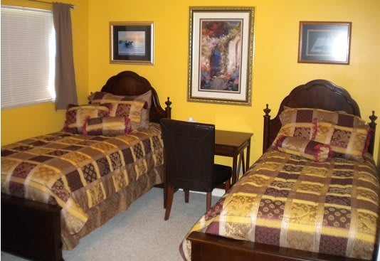 A cozy bedroom with twin beds, adorned with patterned bedding and yellow walls, featuring a desk and artwork on the walls.