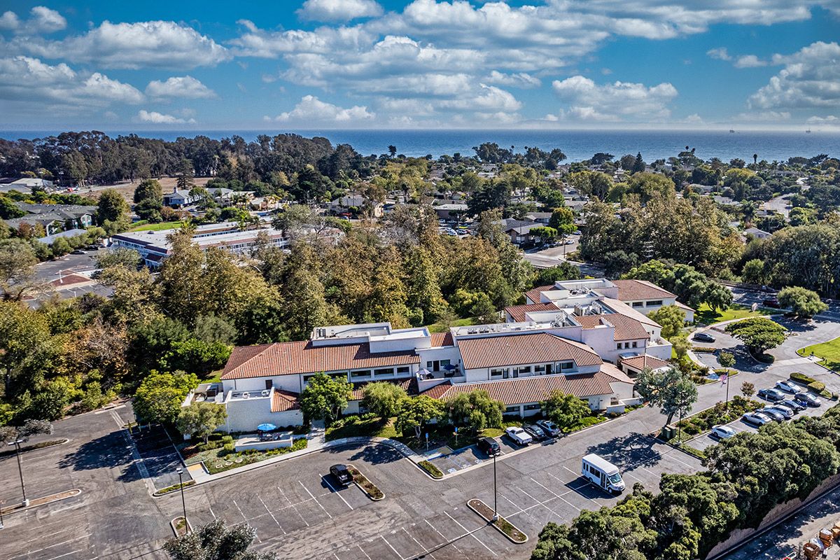 An aerial view of a building surrounded by trees with a parking lot in the foreground and the ocean in the distance.