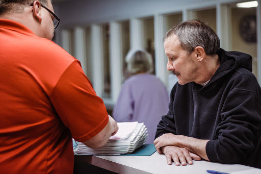 A man engages in a conversation at a service counter, while another person organizes paperwork in the background.