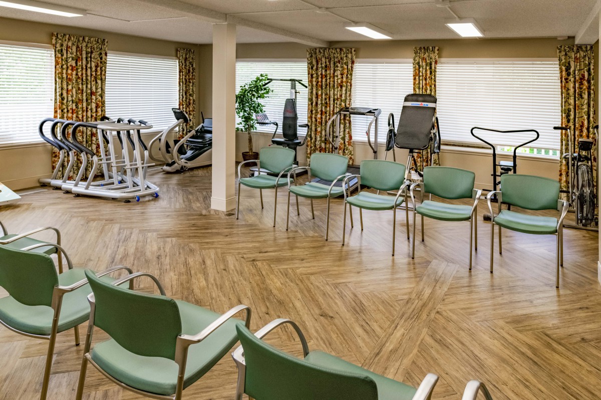 An exercise room featuring fitness equipment and a seating area with green chairs, set on a wooden floor with ample natural light from large windows.