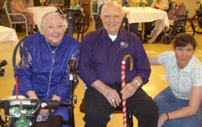 An elderly couple seated indoors; the woman is in a wheelchair, and the man is holding a cane.