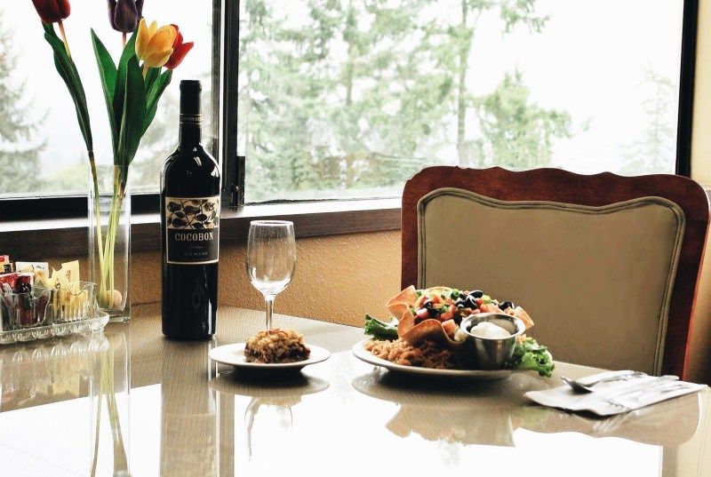 A meal setting featuring a wine bottle, glass, and plates of food on a dining table by the window with flowers in the background.