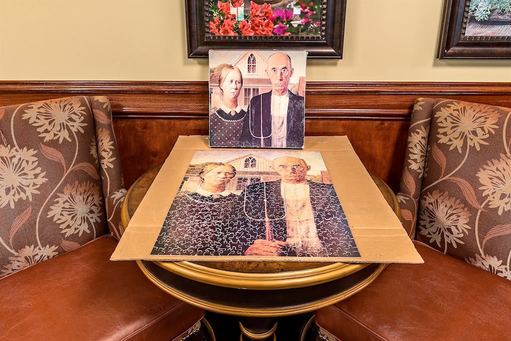 An 'American Gothic' puzzle completed on a round table, with a matching framed artwork displayed above.
