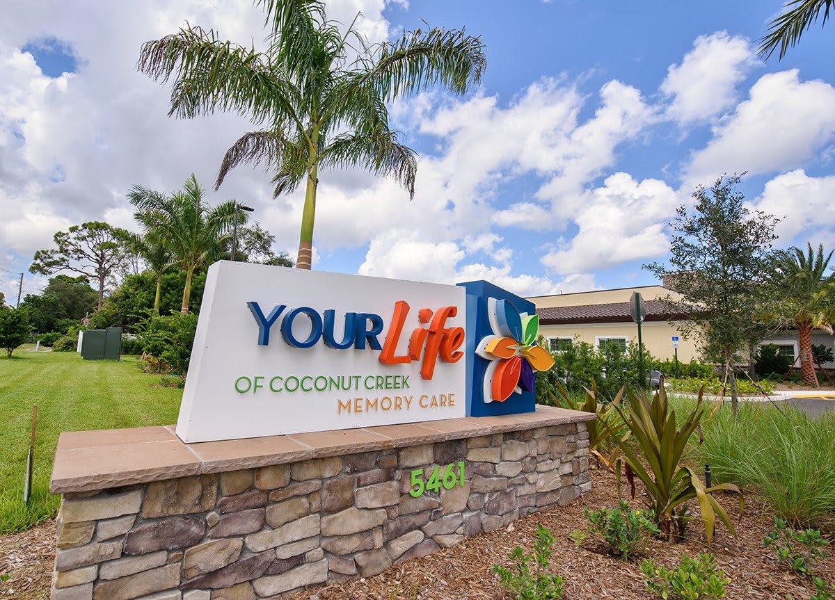 A sign for 'Your Life of Coconut Creek Memory Care' surrounded by palm trees and landscaping.