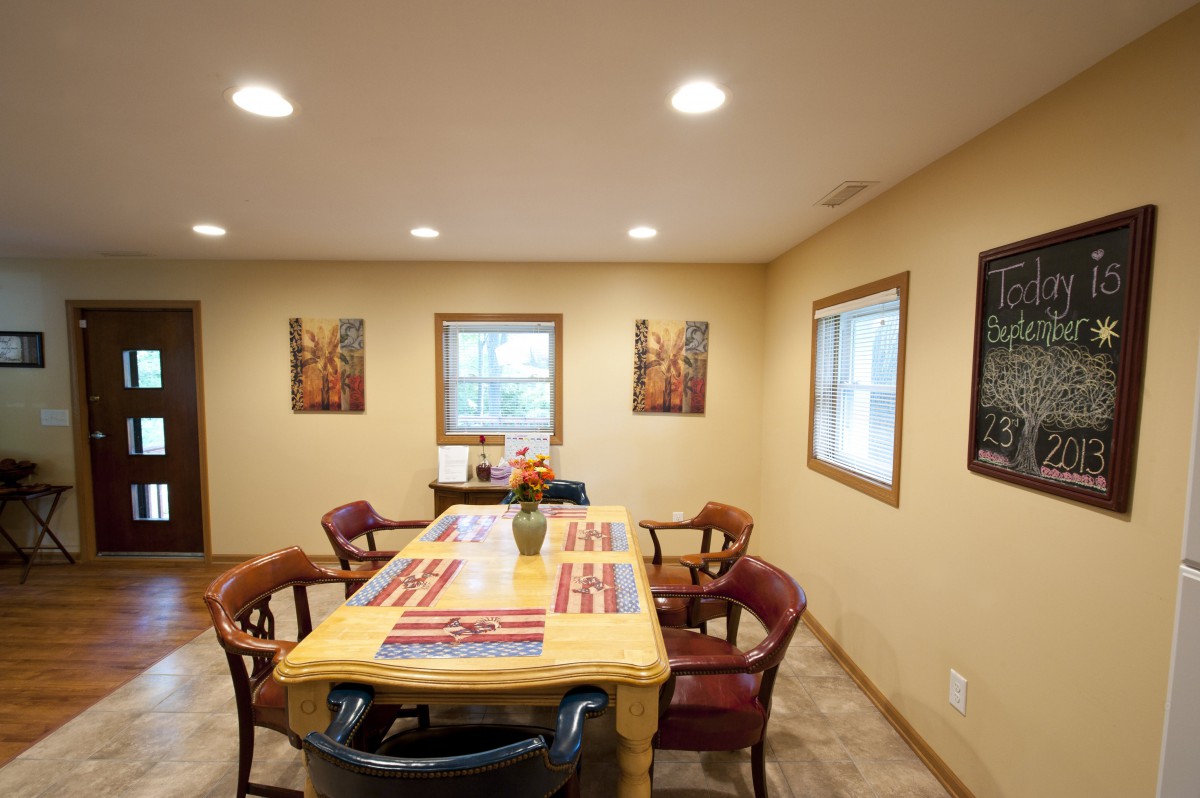 A cozy dining room with a wooden table and chairs, surrounded by artwork and windows providing natural light.