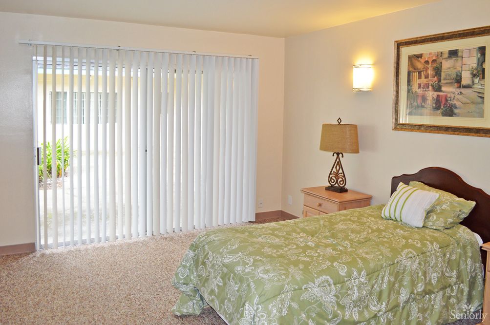 A cozy bedroom featuring a bed with a green patterned cover, a nightstand with a lamp, and vertical blinds covering the window.
