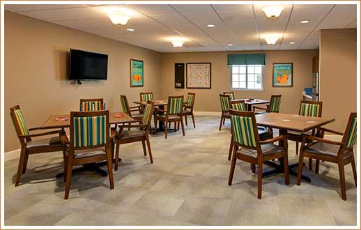 A well-lit community room with several tables and chairs, featuring a television and a window.