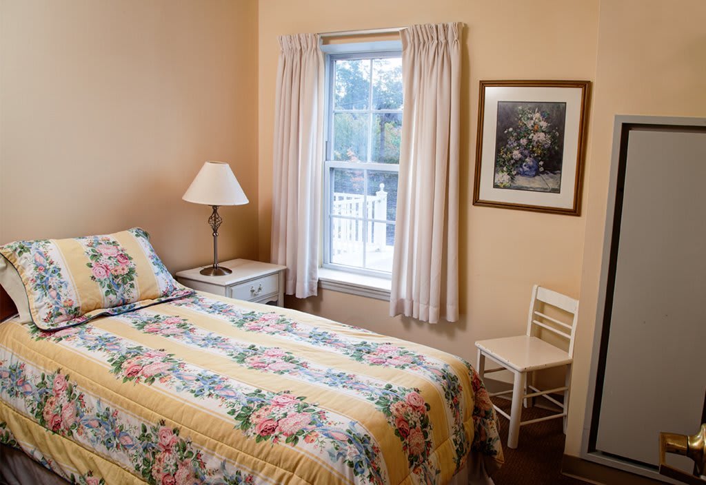 A cozy bedroom with a single bed featuring floral bedding, a lamp on the bedside table, and a window with light curtains.
