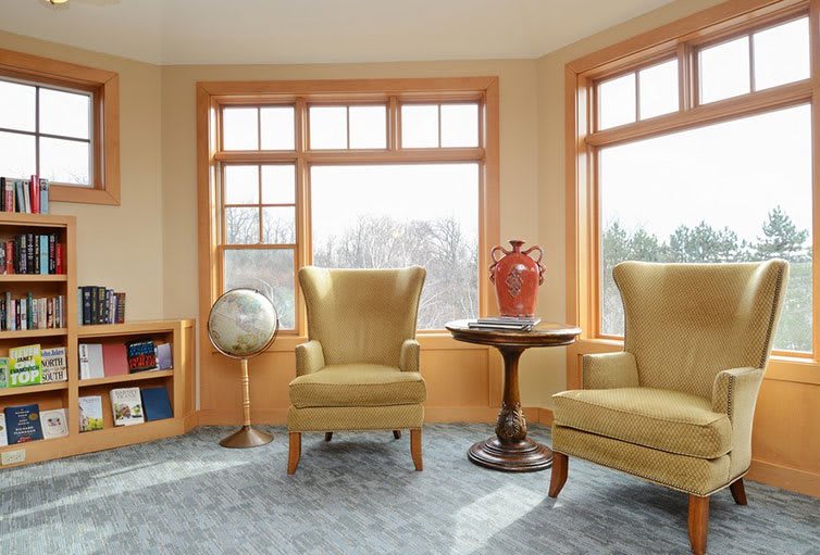 A cozy living room with two armchairs, a small round table holding a red vase, and large windows letting in natural light. A globe sits atop a bookshelf in the corner.