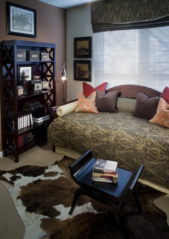 A cozy bedroom featuring a daybed with multiple pillows, a shelf filled with books and decor, and a small table displaying more books.
