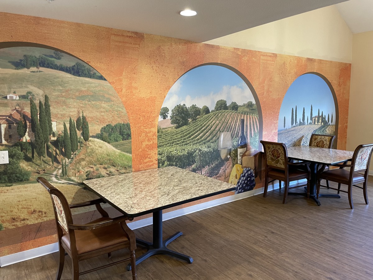 A cozy restaurant interior featuring a scenic mural depicting a countryside landscape, with tables and chairs arranged for dining.
