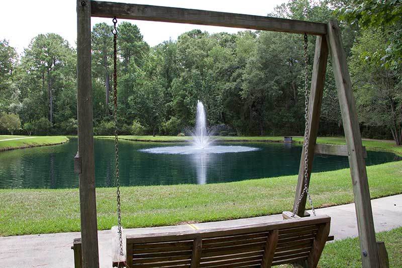 A peaceful scene with a wooden swing overlooking a pond and water fountain surrounded by trees.