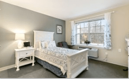 A cozy bedroom with a white bed, nightstand, and a large window allowing natural light to fill the room.