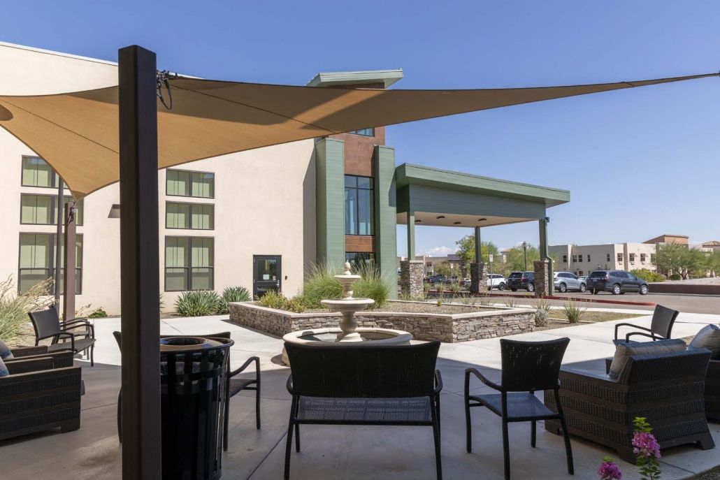 A modern outdoor patio area with shade sails, seating, and a decorative fountain in front of a contemporary building.