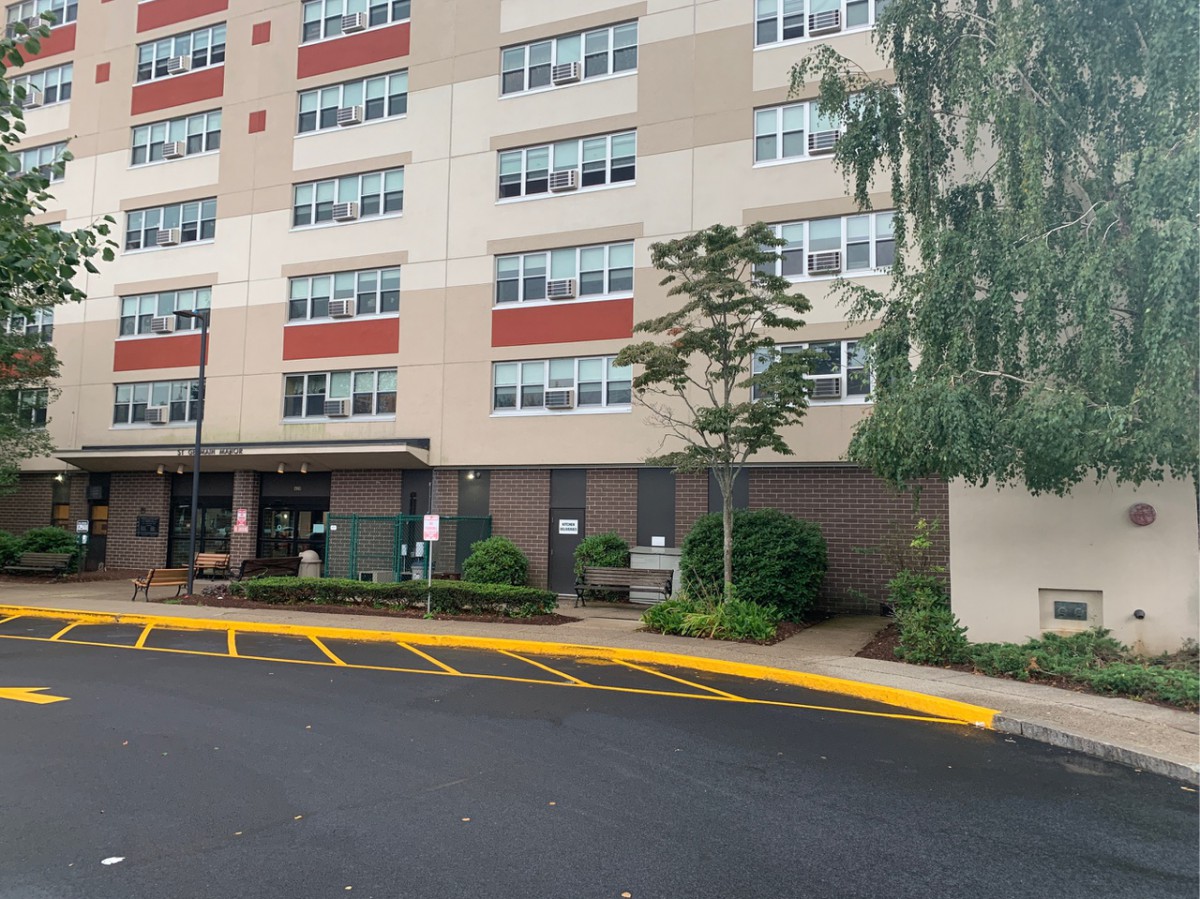 Exterior view of a residential apartment building entrance with surrounding greenery and parking area.