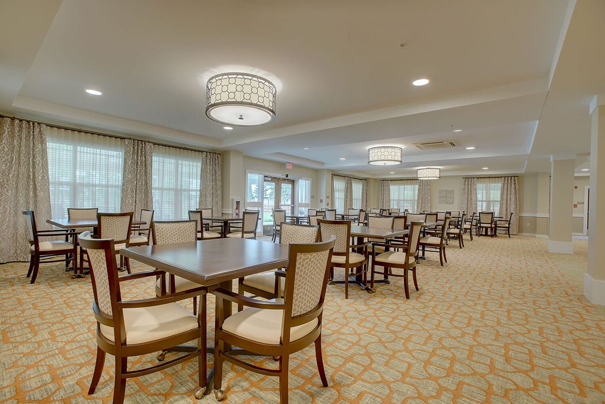 A spacious dining room with multiple tables and chairs, featuring elegant chandeliers and natural light from large windows.