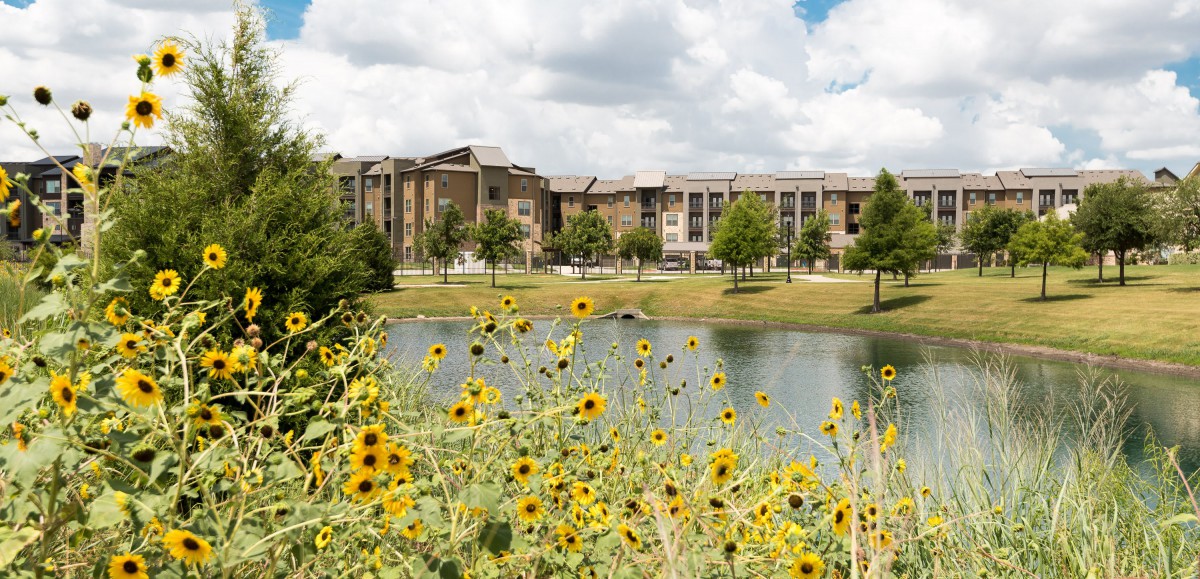 A scenic view of a building by a lake with wildflowers in the foreground and a partly cloudy sky.