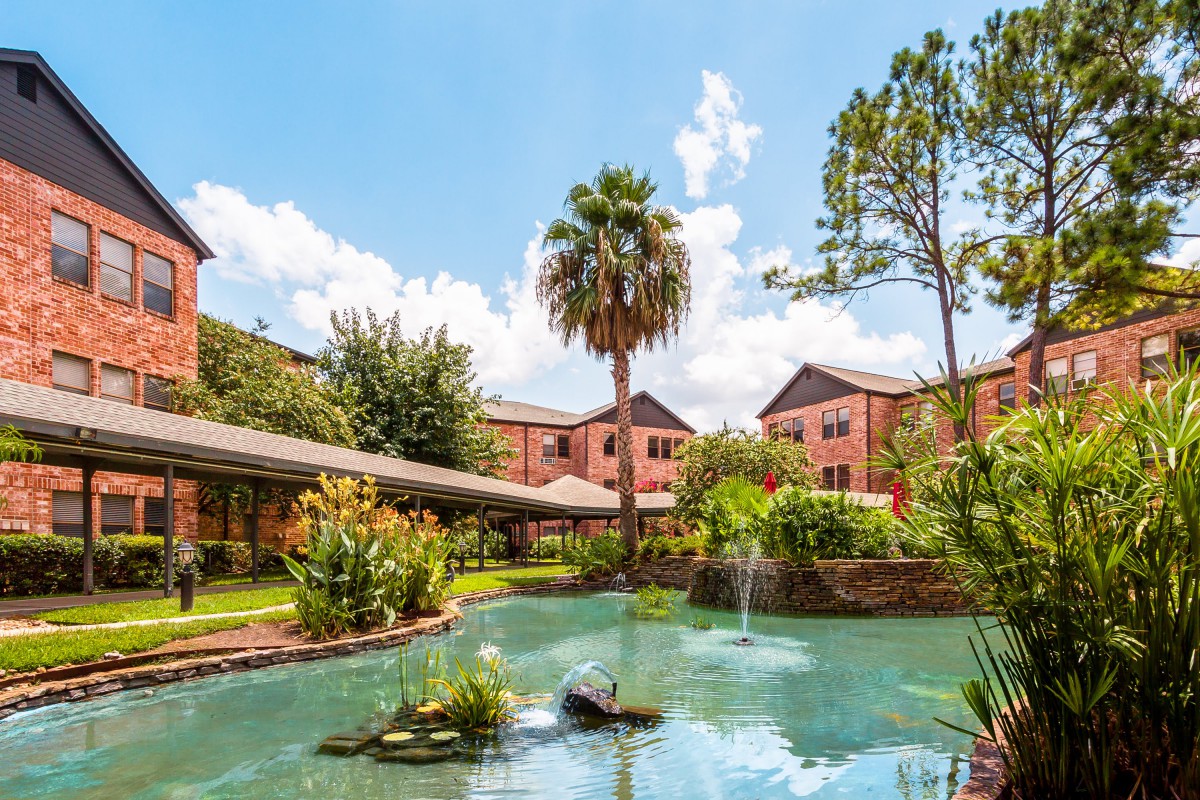 A serene view of a garden with a pond and fountain, surrounded by lush greenery and residential buildings under a bright blue sky.