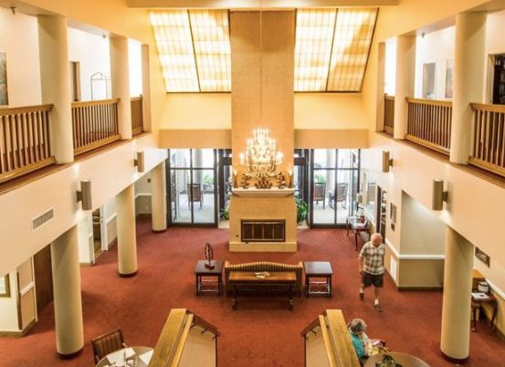 A spacious lobby area with a chandelier, carpeted flooring, and various furniture pieces, including tables and chairs. People are seen walking around the space.