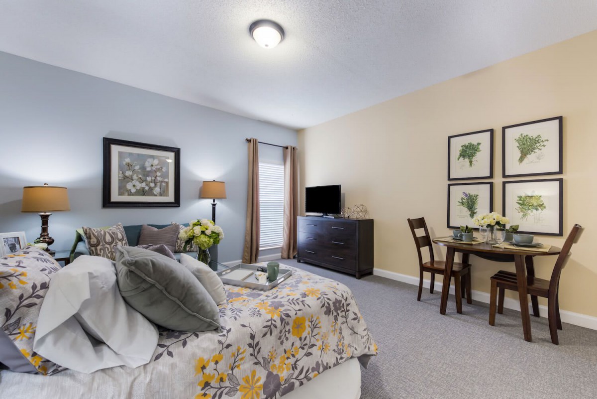 A cozy bedroom interior with a small dining area, decorated with floral bedding and artwork.