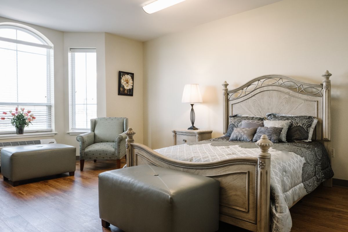 A cozy bedroom featuring a large bed with a decorative headboard, accompanied by two chairs and a lamp, bathed in natural light from a tall window.