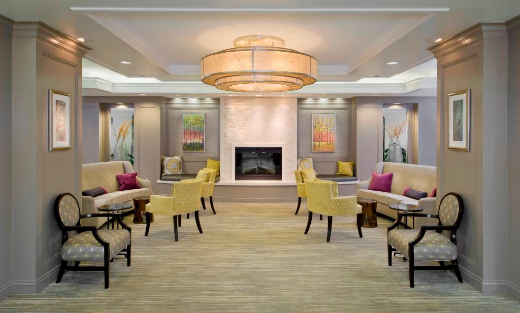A modern lobby with a central fireplace, surrounded by seating and artwork, featuring a large chandelier.