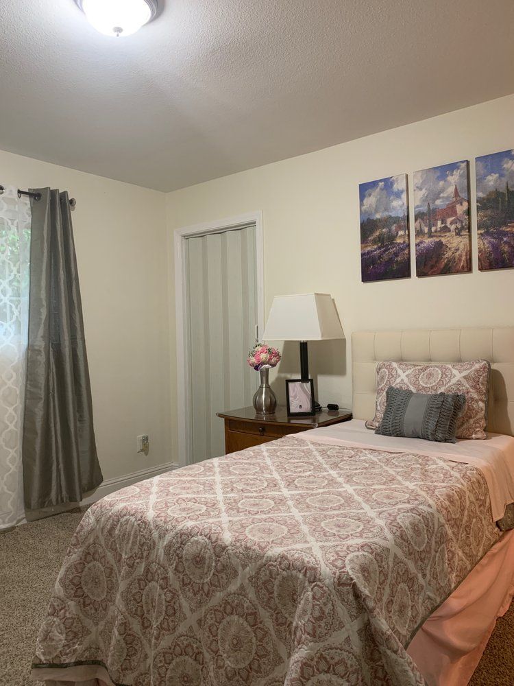 A cozy bedroom with a single bed, adorned with patterned bedding and a decorative throw pillow. The room features curtains, a bedside table with a lamp, floral arrangement, and wall art above the headboard.
