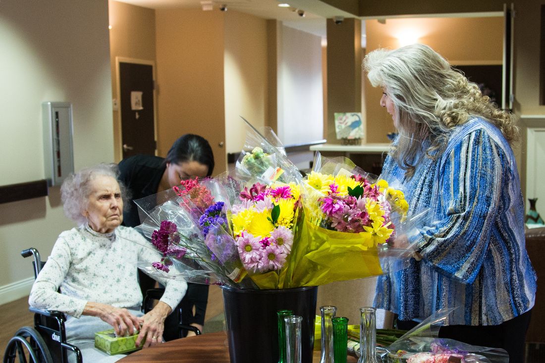 Elderly woman receiving visitors in a nursing home with vibrant bouquets of flowers around.