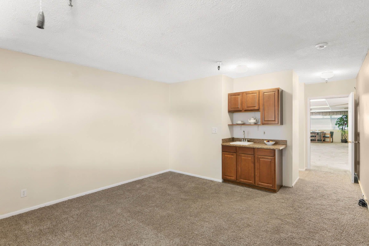 An empty studio apartment with a small kitchenette featuring wooden cabinets and a sink.