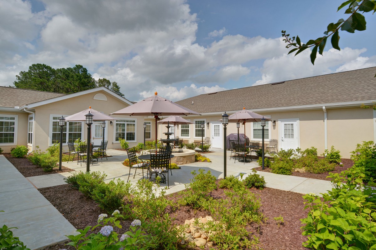 A well-maintained outdoor patio area with tables and umbrellas, surrounded by landscaping, adjacent to a building under a partly cloudy sky.