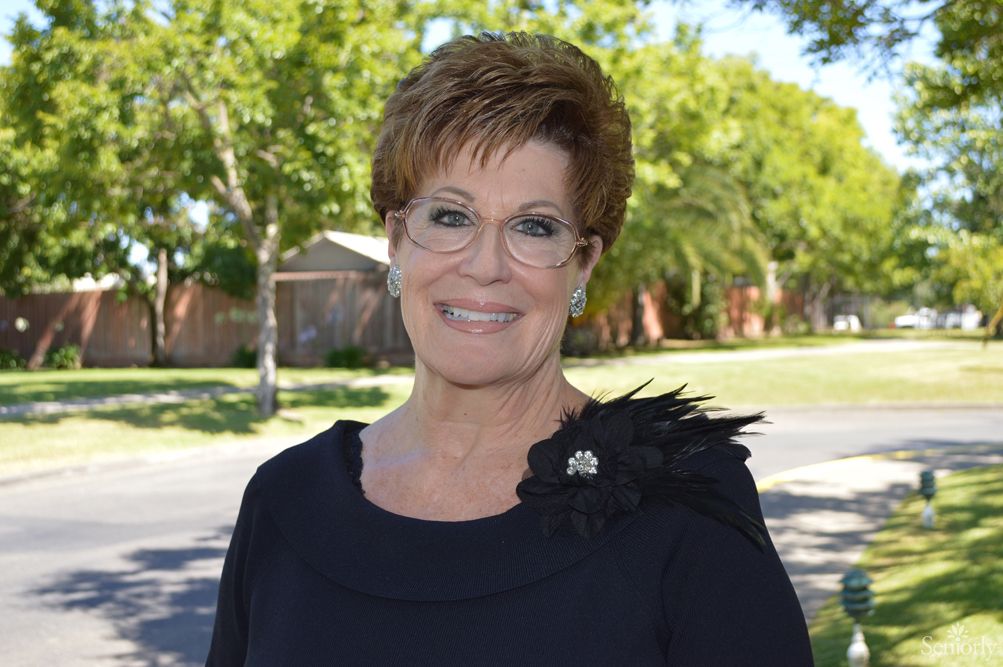 A woman with glasses smiling in an outdoor setting with trees in the background.