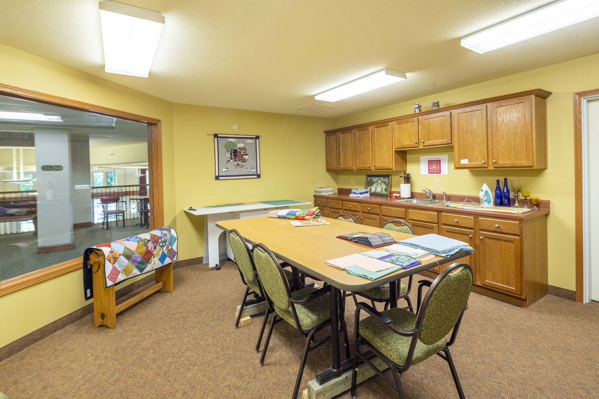 A well-lit craft room with a large table surrounded by chairs, featuring quilting supplies and storage cabinets.
