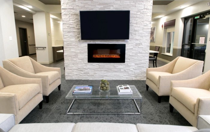 A modern living room with a mounted TV and fireplace, featuring a glass coffee table between two beige armchairs.