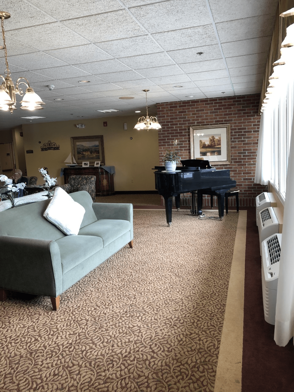 A cozy living room with a sofa and grand piano, featuring carpet flooring, brick walls, and large windows.