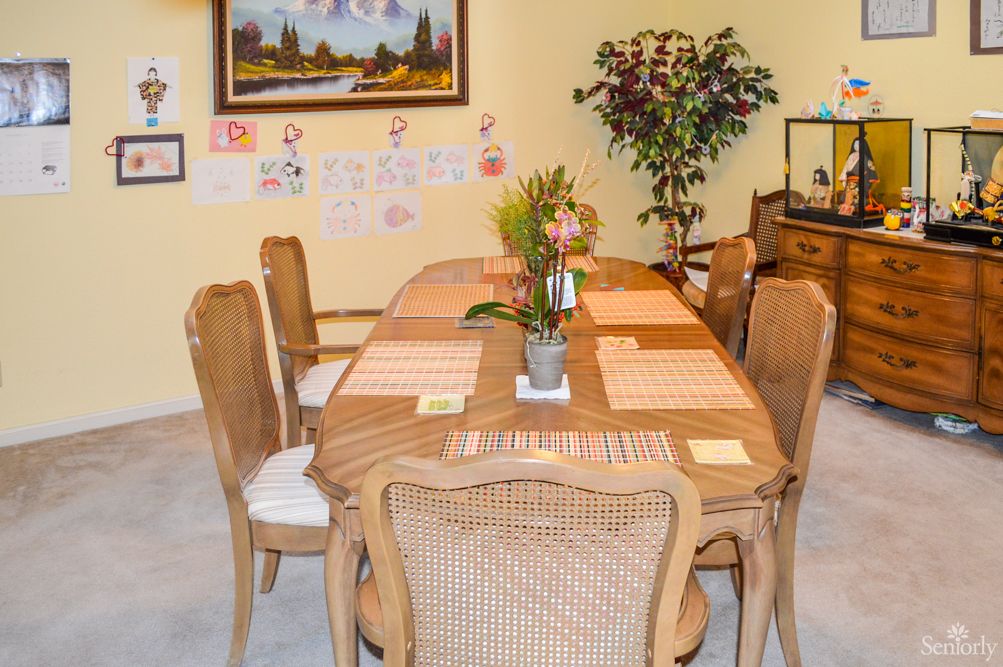 A cozy dining room with a wooden table set for six, adorned with placemats and a centerpiece, surrounded by chairs and decorated with wall art and plants.