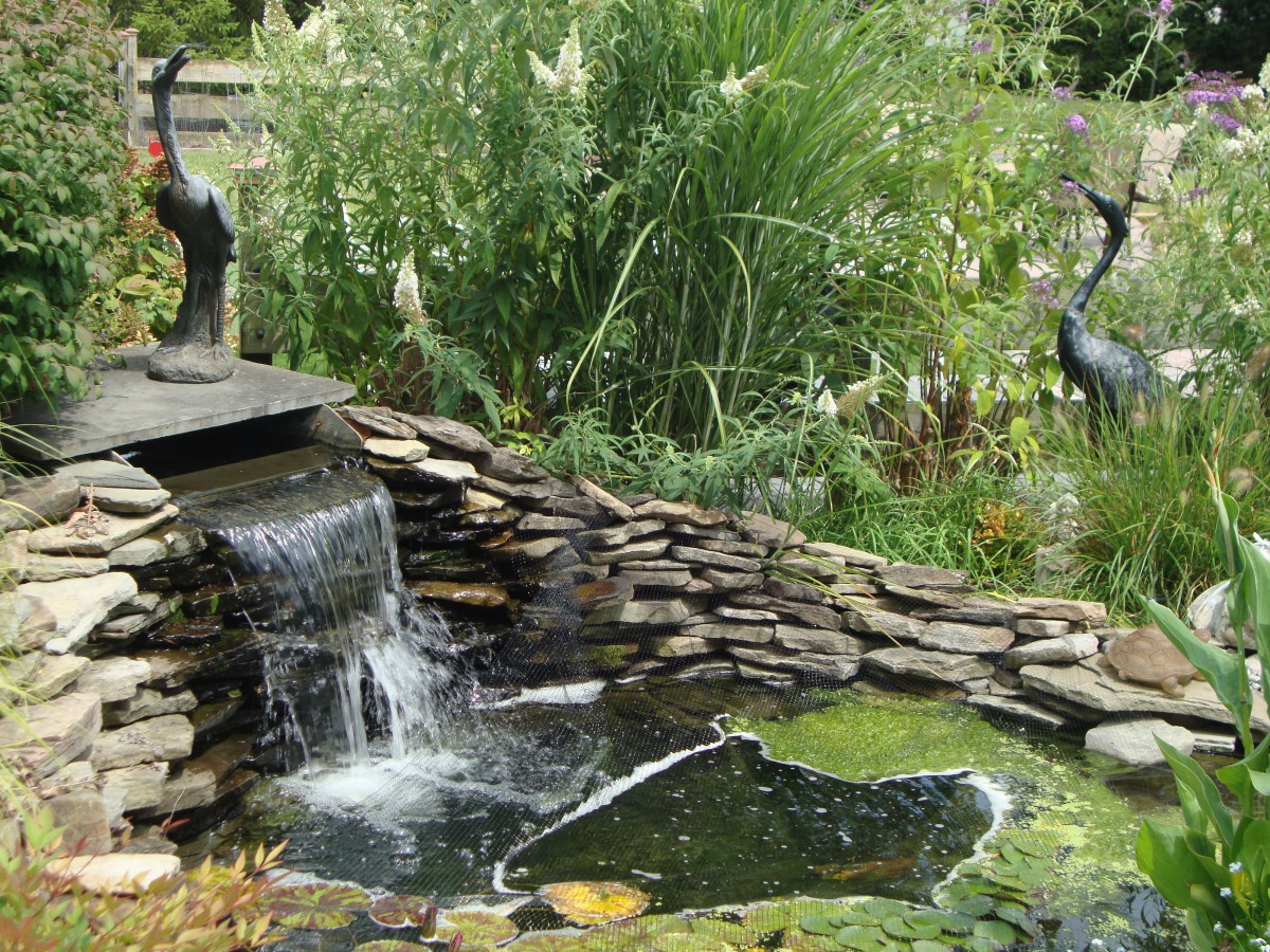 A serene garden pond with a small waterfall and sculptures surrounded by lush greenery.