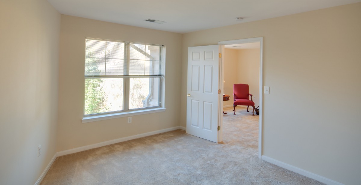 A bright, empty room with large windows and beige carpet, featuring an adjacent room with a red chair visible through an open door.