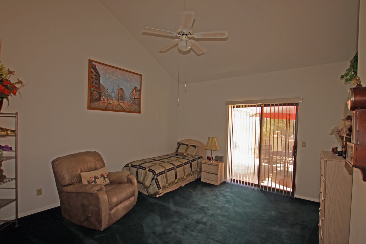 A minimalist bedroom with a single bed, an armchair, and a nightstand, featuring a ceiling fan and large window with blinds.